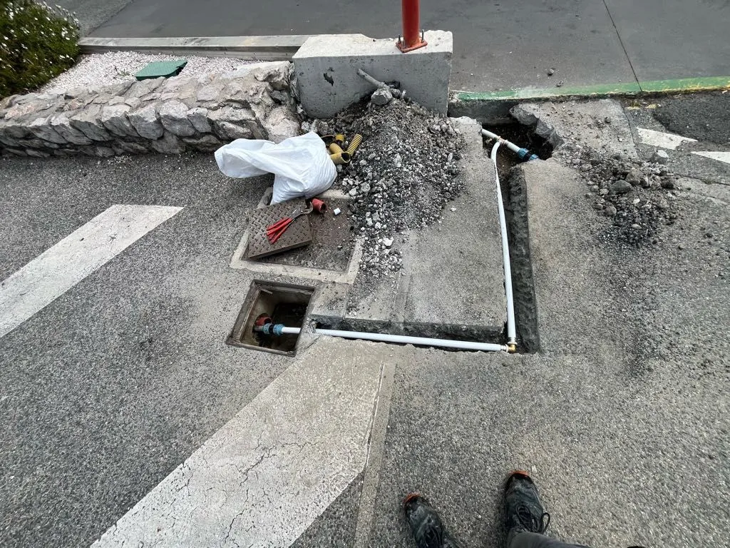 Tranchée ouverte dans un trottoir et un parking, avec regard d'eau ouvert, avant réparation